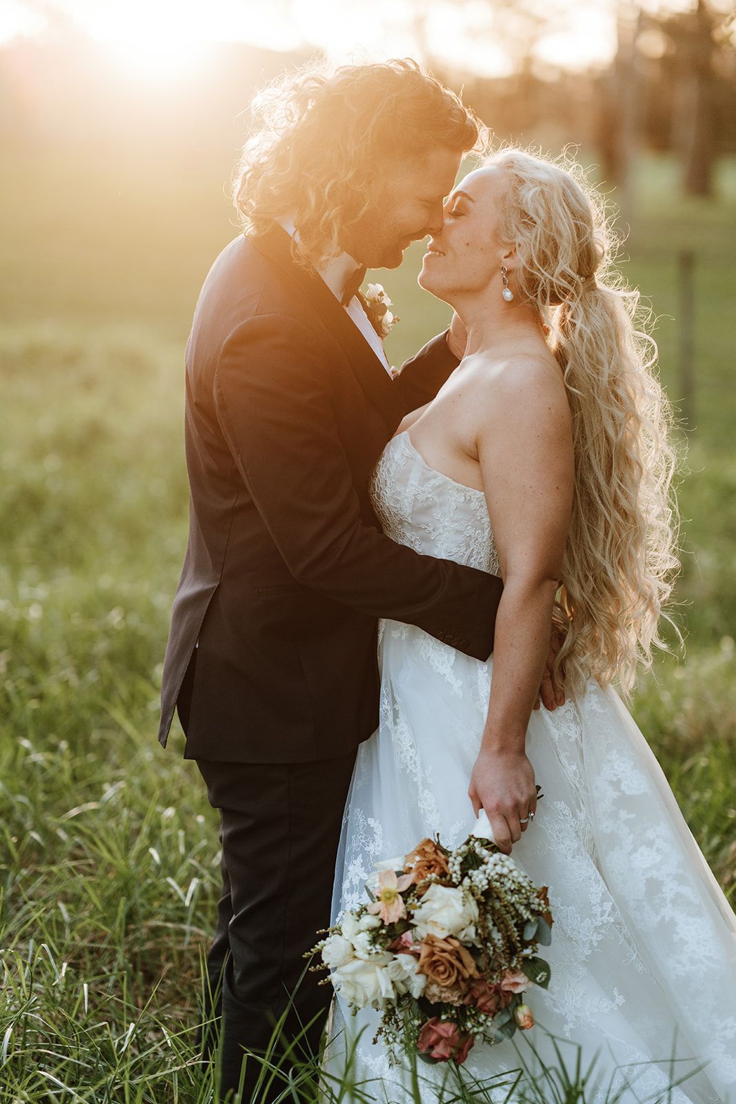 Couple kissing in a field at sunset; woman in white dress, man in suit, holding bouquet.