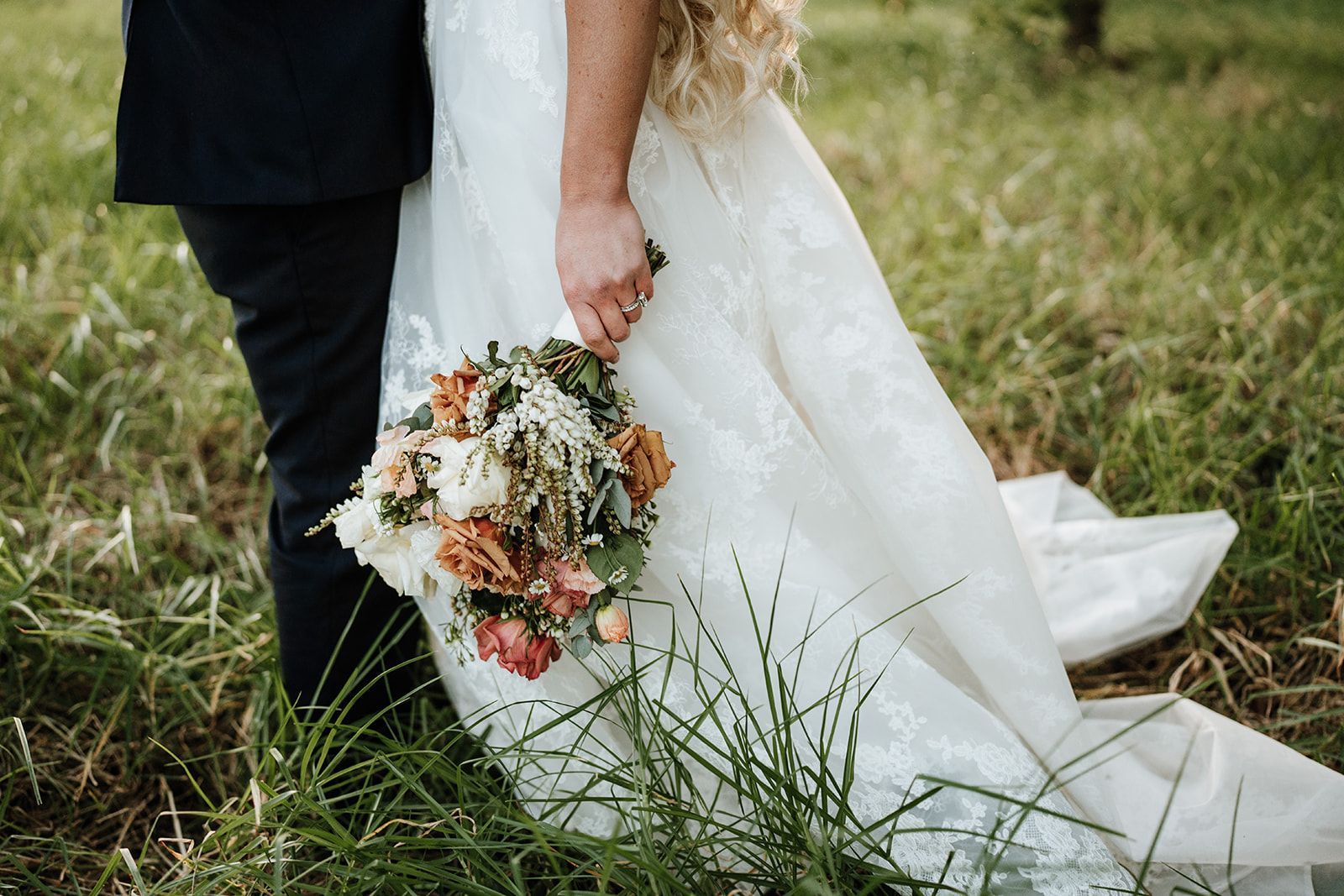 Bride and groom holding hands, wedding bouquet in tall grass.