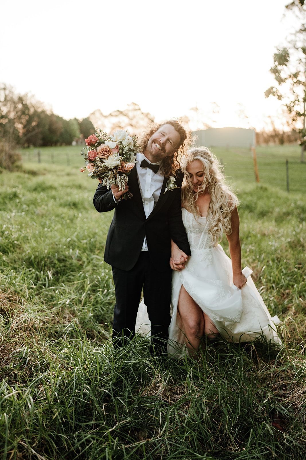 Newlyweds holding hands in a field, groom smiling with bouquet, bride looking down. Golden hour light.