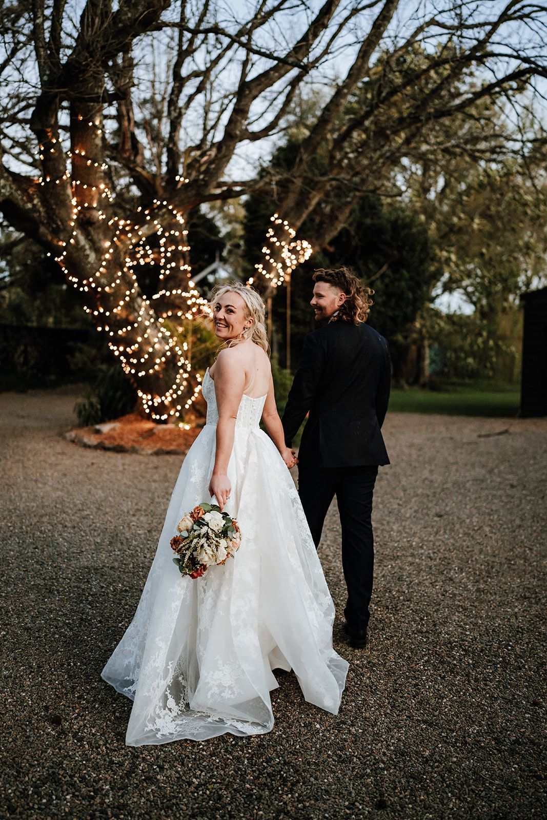 Bride and groom holding hands, smiling. Bride in white dress, groom in black suit, near tree with lights.
