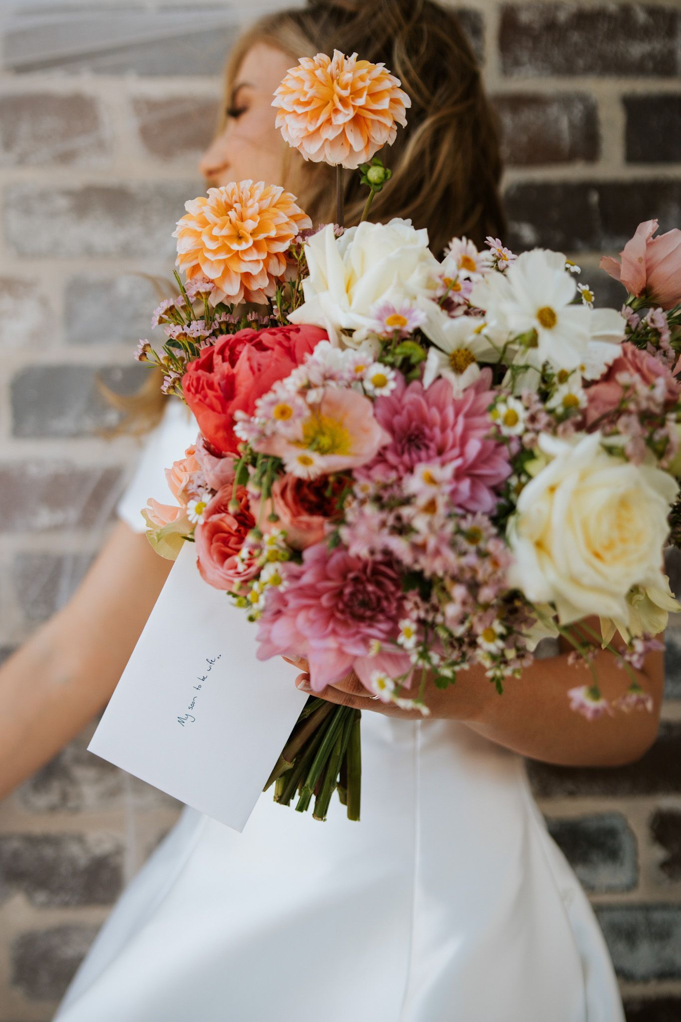 Woman holding a vibrant bouquet of flowers, standing against a brick wall. She wears a white dress.