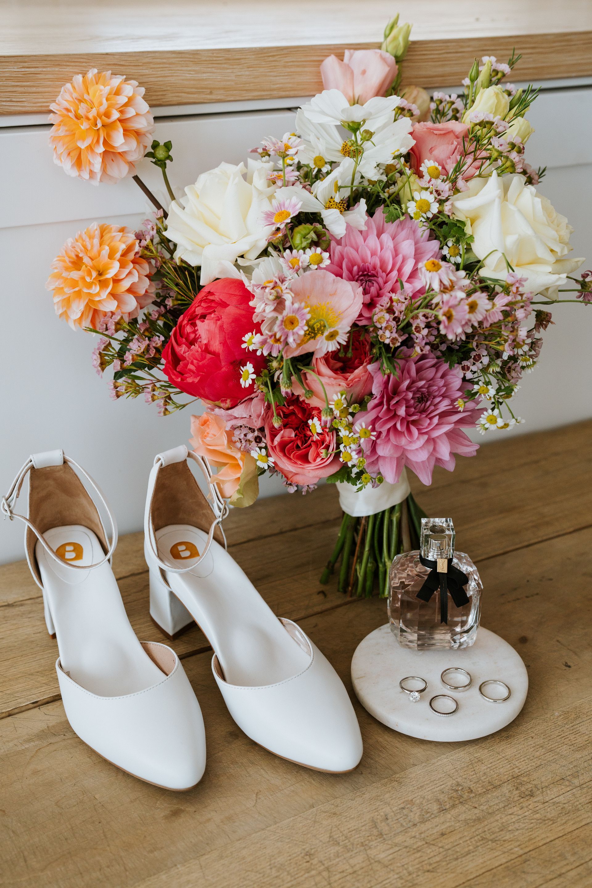 Wedding bouquet with shoes, perfume, and rings arranged on wood.