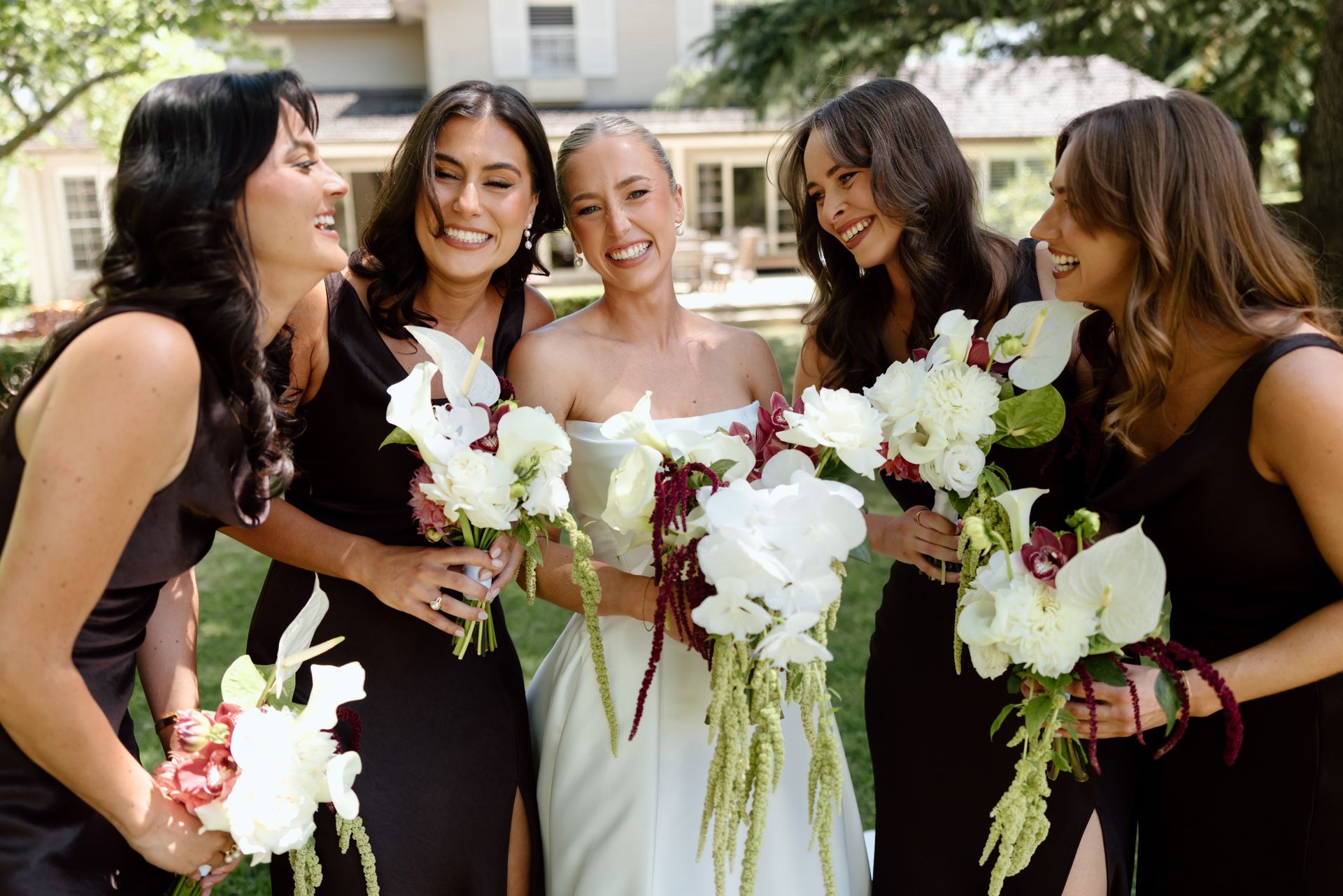 Bride and four bridesmaids in black dresses holding white bouquets outdoors, smiling and laughing