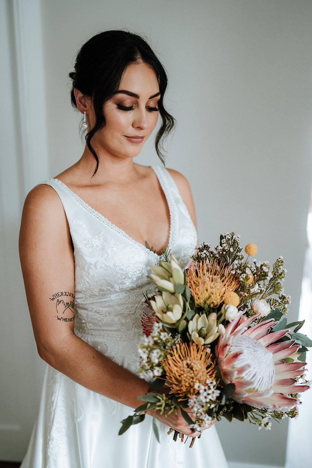 Bride in white gown holding bouquet, looking down, indoor setting.