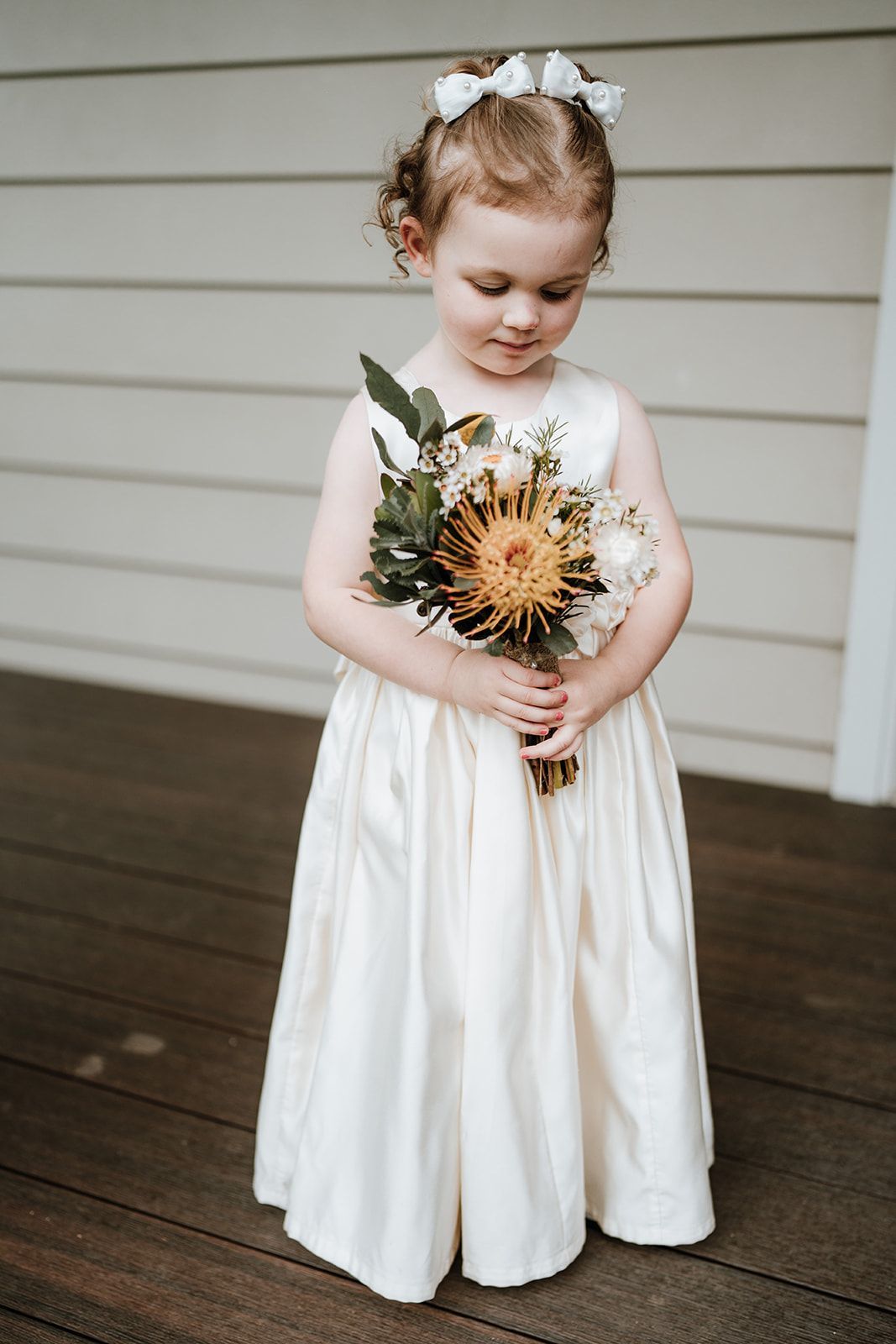 Young girl in a cream dress holds a bouquet, standing on a porch. She has curly hair and a bow.