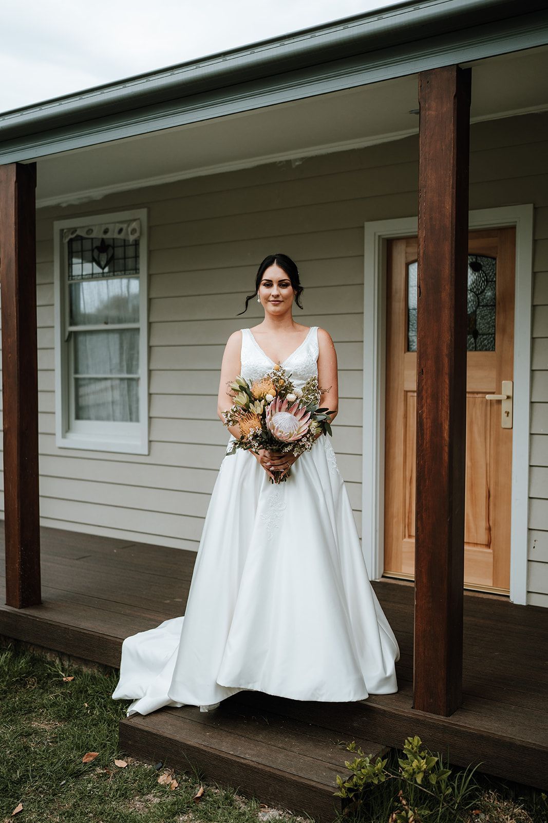 Bride in white wedding dress holding flowers, standing on porch with wood columns.