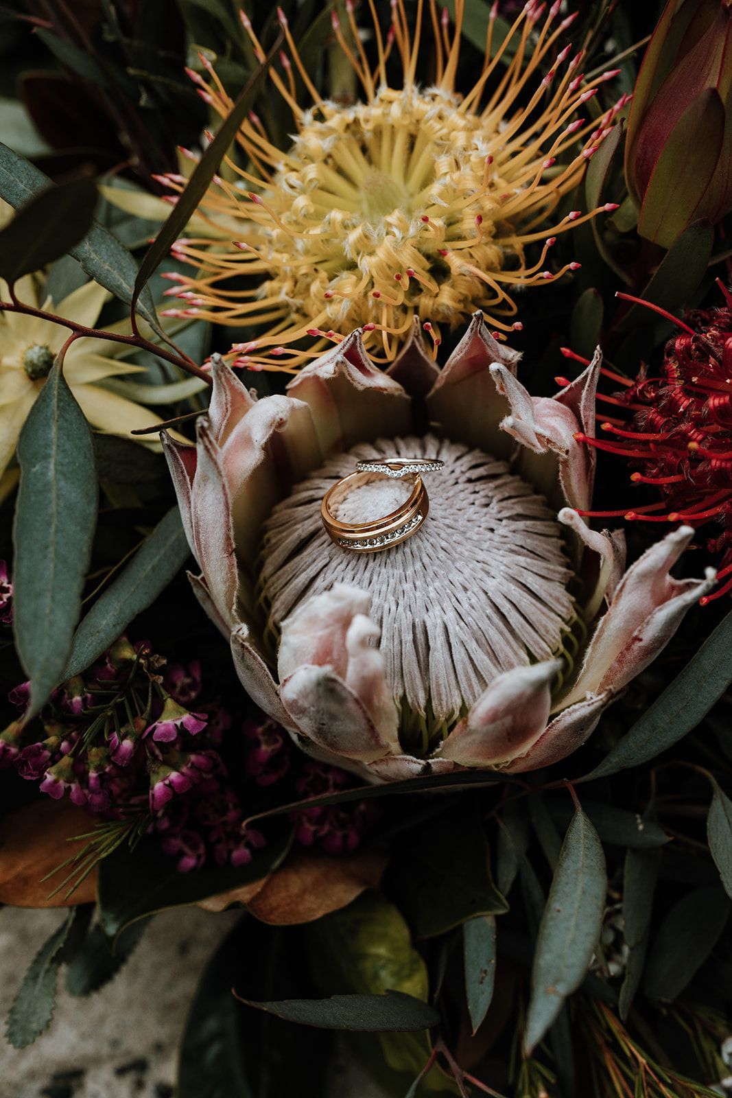 Wedding rings on a protea flower in a bouquet with yellow, red, and green foliage.