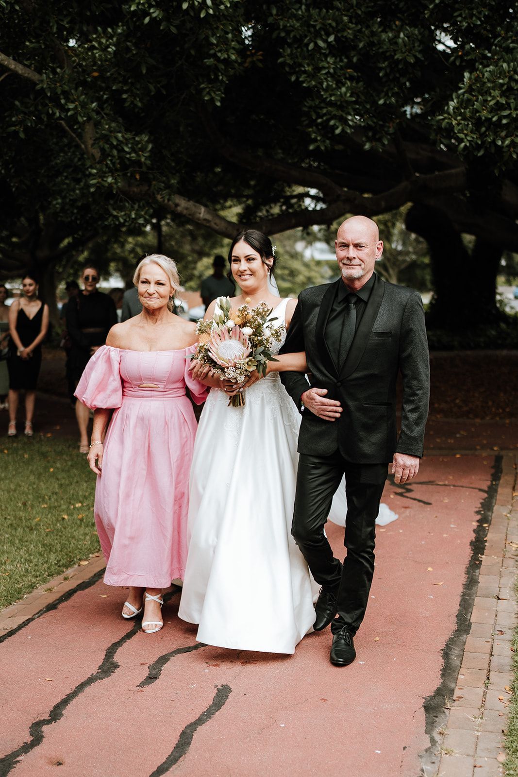 Bride walks down aisle with parent on each side; pink dress, black suit.