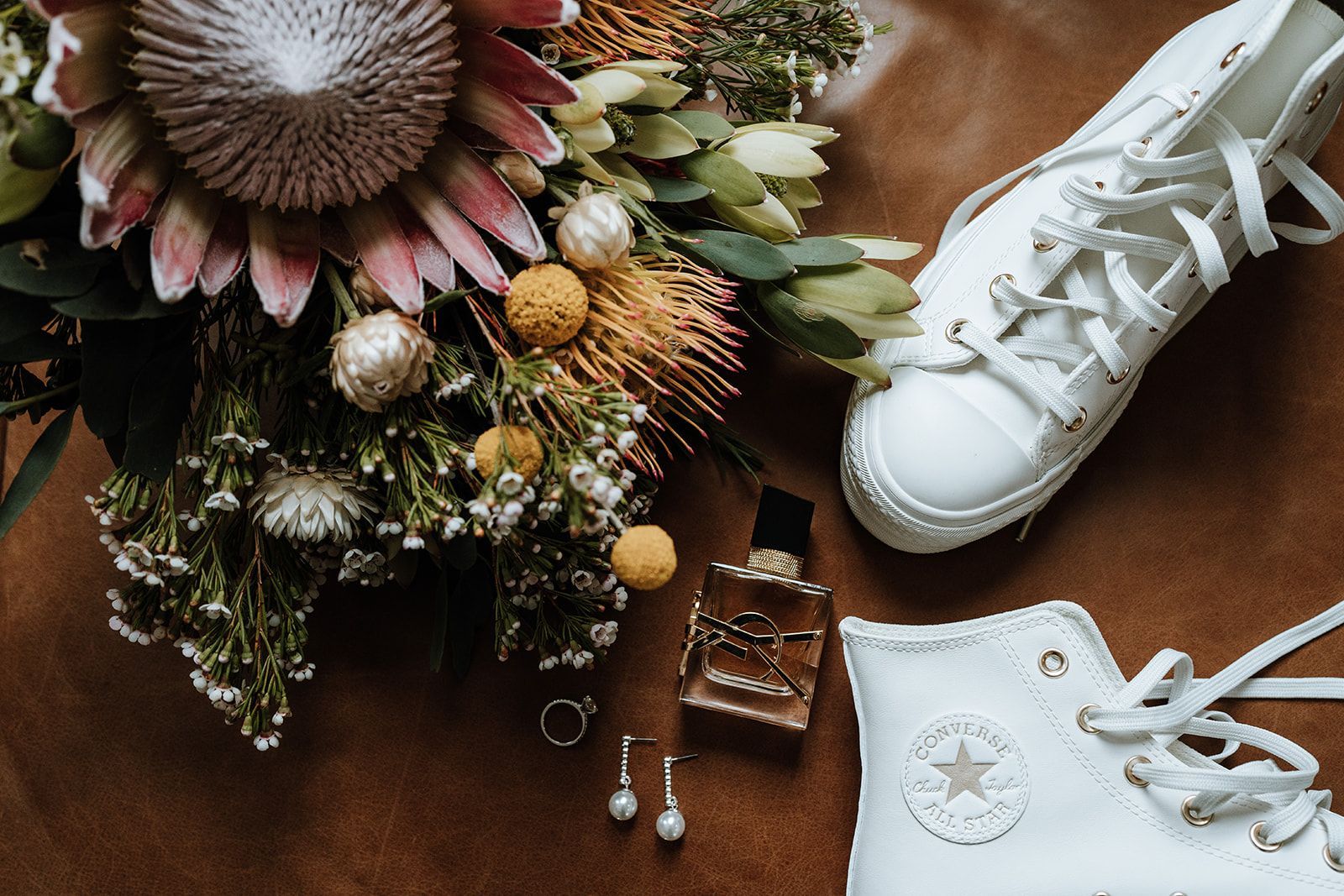 Wedding details: bouquet, white Converse sneakers, perfume bottle, jewelry on brown surface.