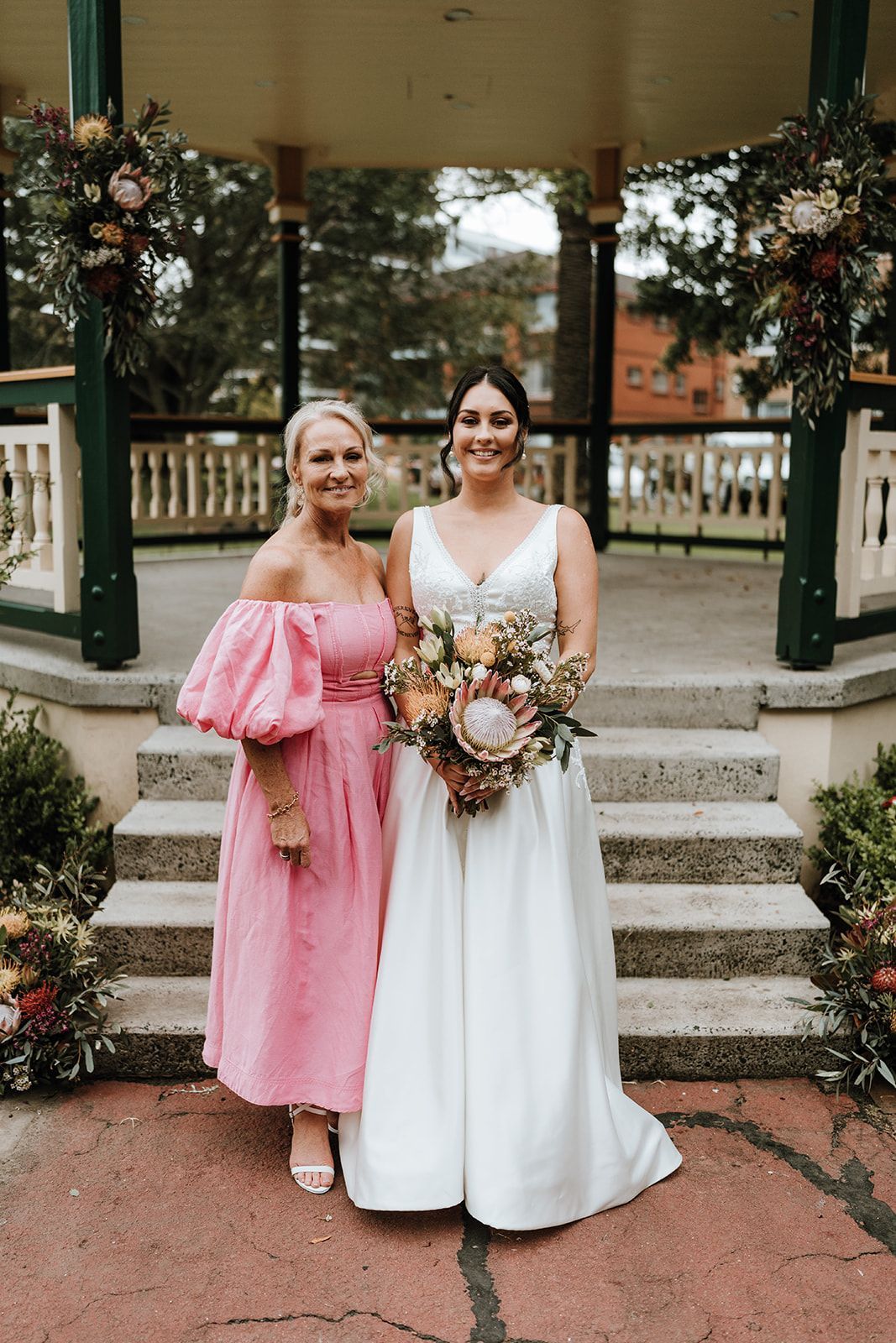 Bride and woman in pink dress pose in front of a gazebo, holding flowers.