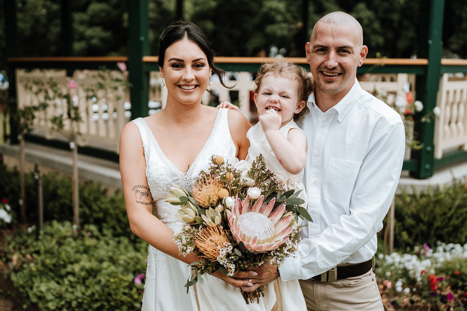 Family of three smiling, posing outdoors. Woman in white dress, man in white shirt, and a child holding flowers.