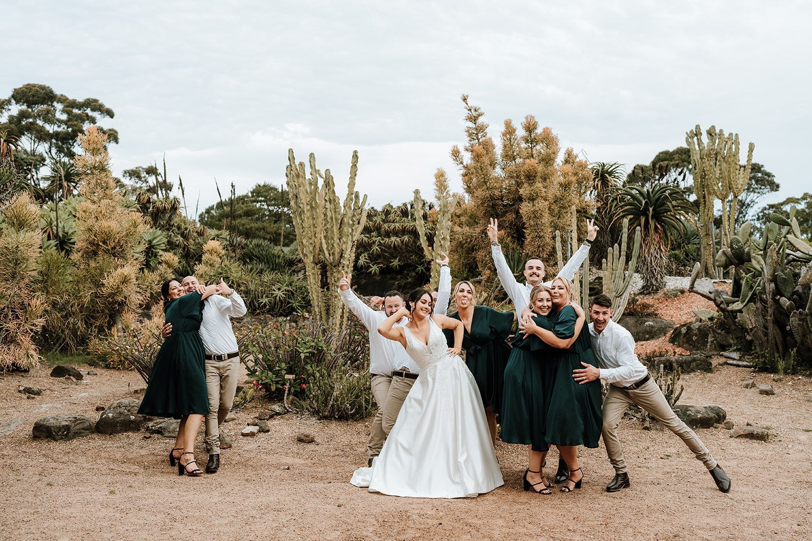 Wedding party posing in a botanical garden; bride in white gown, bridesmaids in green, others in tan pants and white shirts.