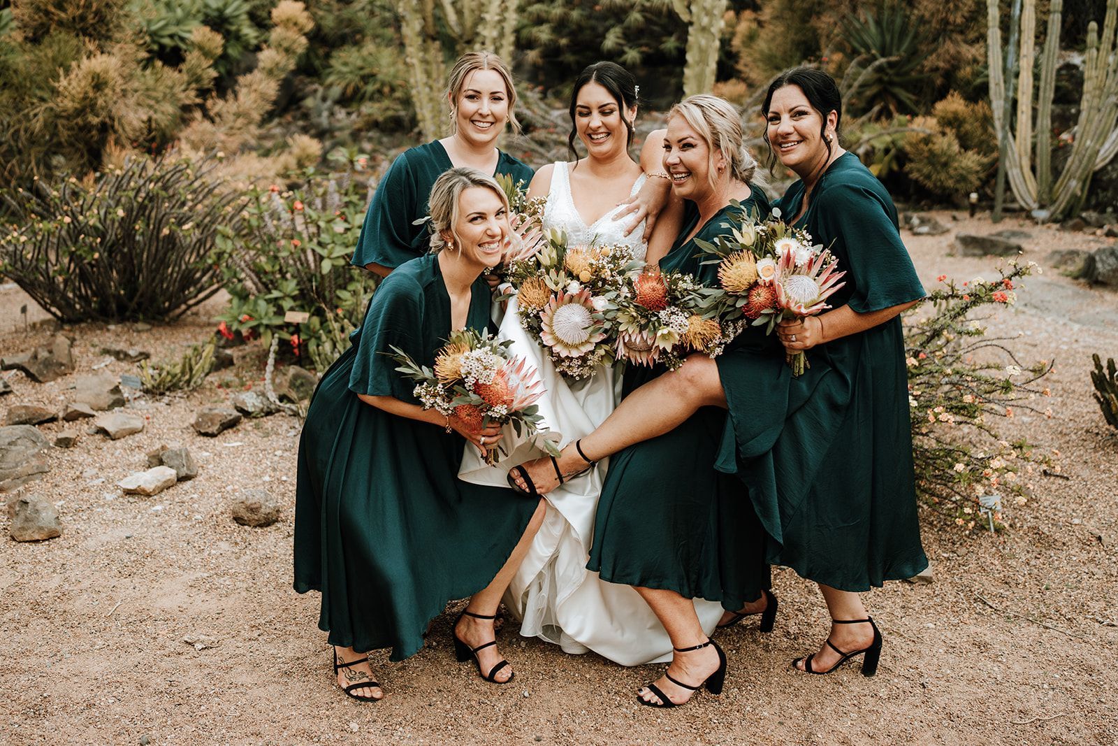Bride poses with bridesmaids in a garden, all smiling. They wear teal dresses and hold bouquets.