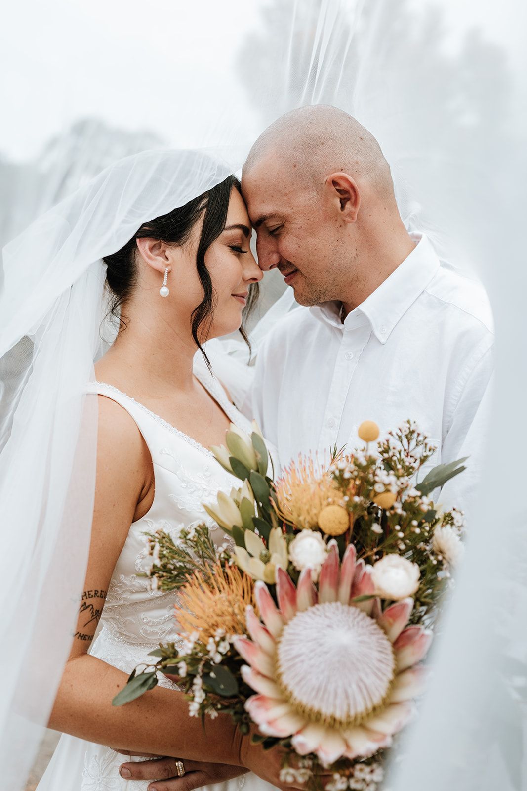 Bride and groom touch foreheads, holding flowers. She wears a veil and dress, he wears a white shirt. Outdoors.