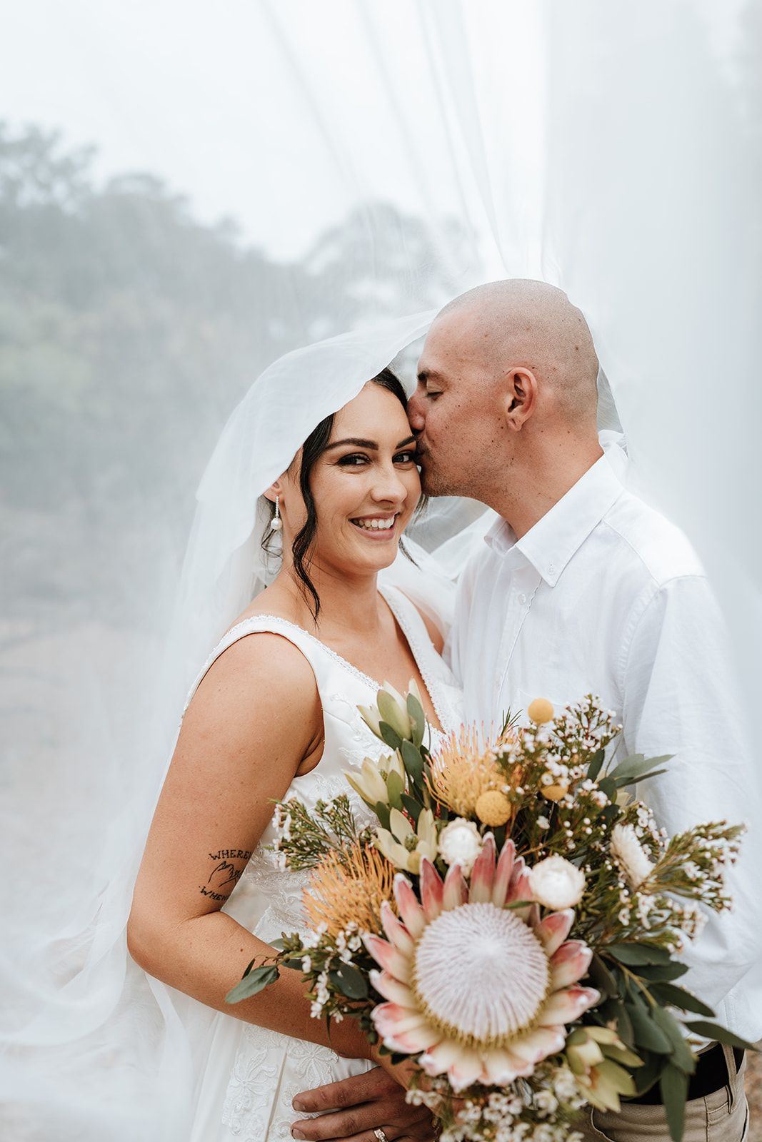 Bride and groom embrace, he kisses her forehead. She smiles, holding a bouquet, under a veil. Outdoors, trees in background.