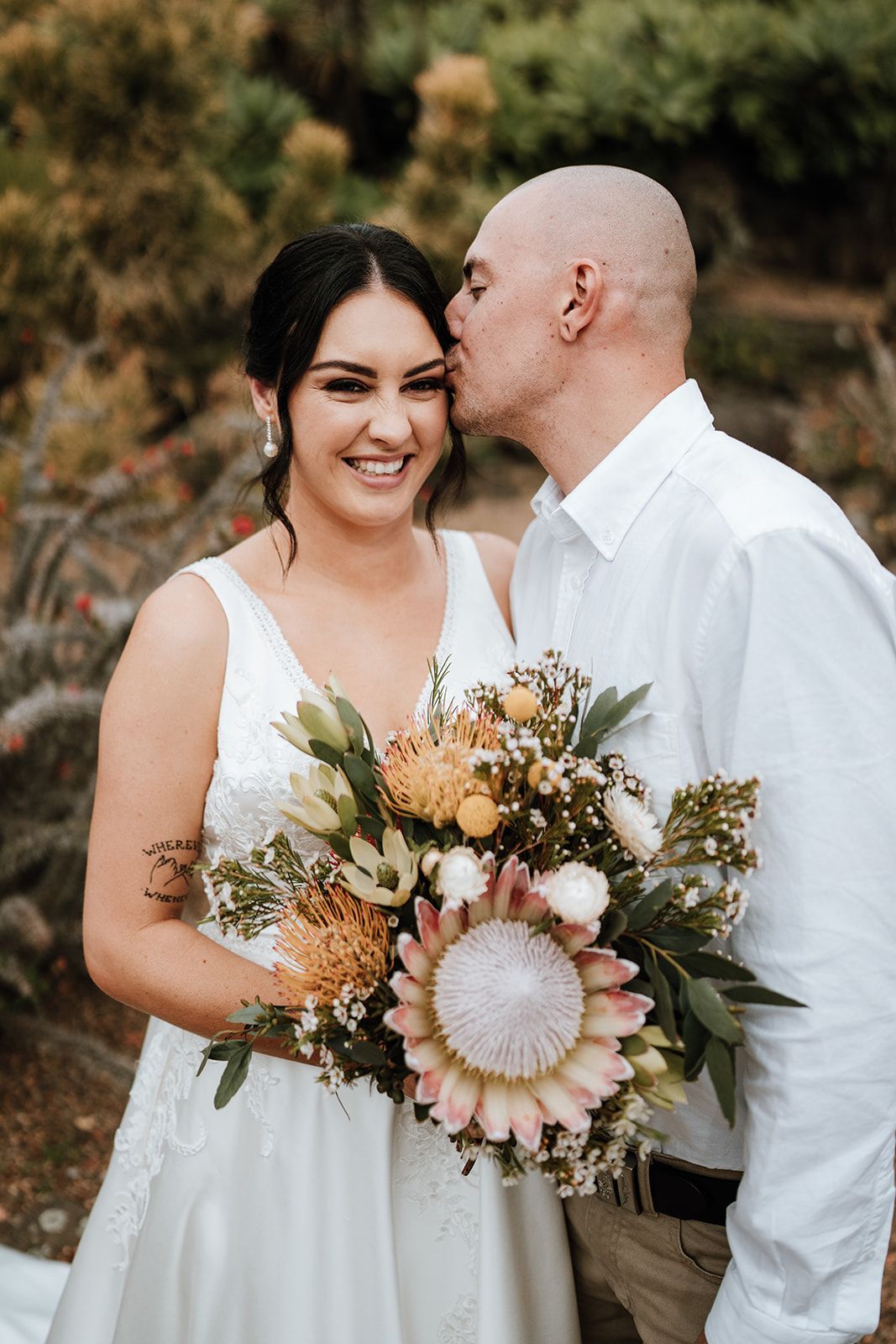 Bride and groom smiling outdoors; man kisses woman's forehead; woman holds bouquet of flowers.