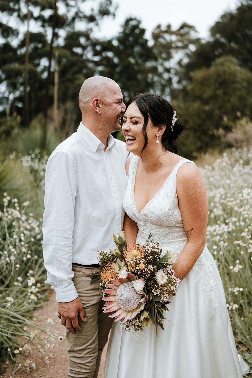 Bride and groom laughing in a field. Bride in white dress, holding bouquet. Groom in white shirt, khaki pants.
