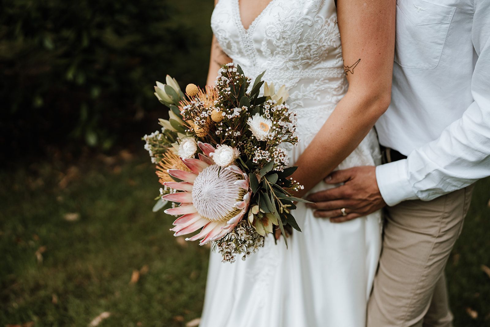 Bride in white dress holding bouquet, groom's hand on her waist. Outdoor setting, green foliage.