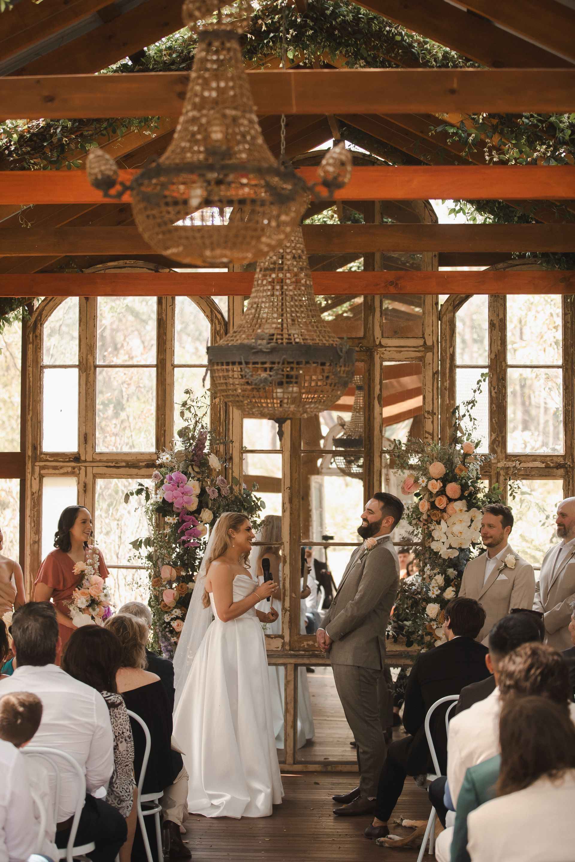 Bride and groom at altar during wedding ceremony. Floral arrangements, chandelier, wooden beams, guests seated.