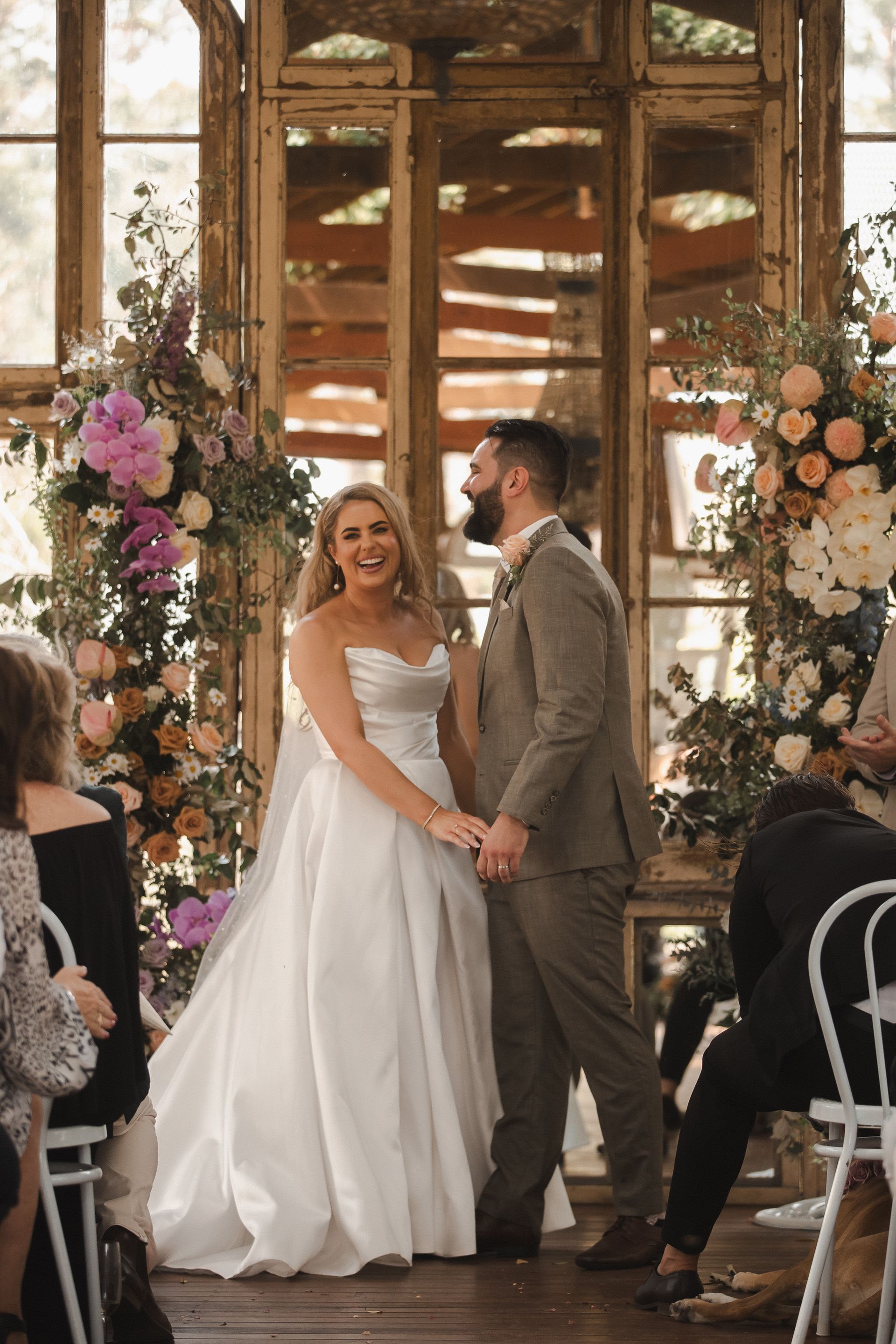 Wedding ceremony with bride in white dress and groom in suit, holding hands, smiling, in front of mirrored backdrop.