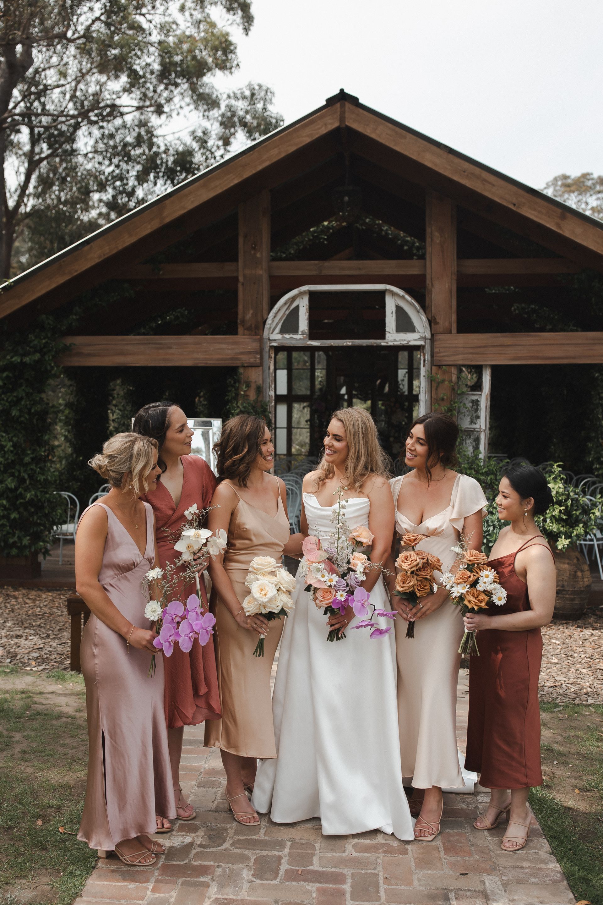 Bride and bridesmaids pose outside a wooden building. They hold bouquets, wearing colorful gowns, smiling.