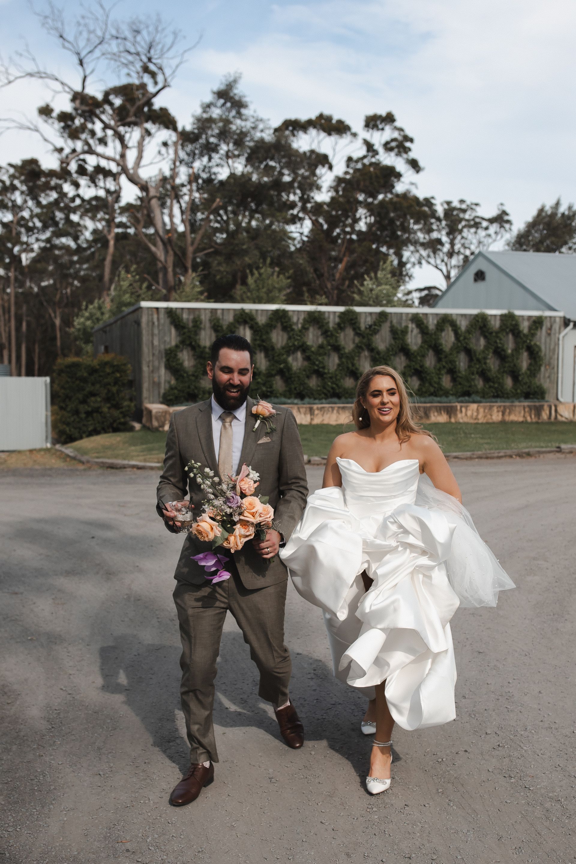 Couple in wedding attire walking outdoors; bride lifts dress, groom holds bouquet.