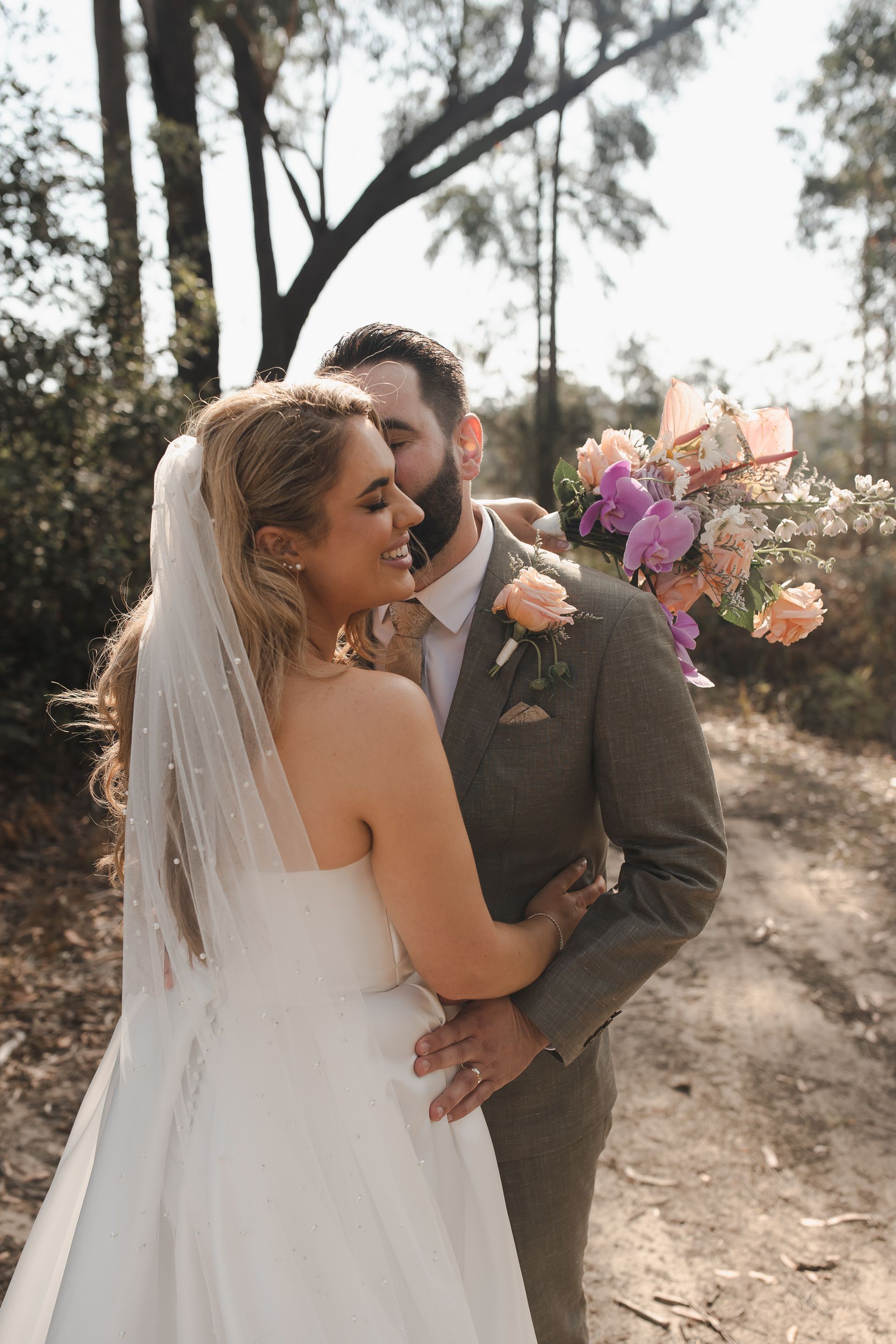 Bride and groom embrace outdoors; she wears a veil and white gown, he wears a tweed suit, sunlight.