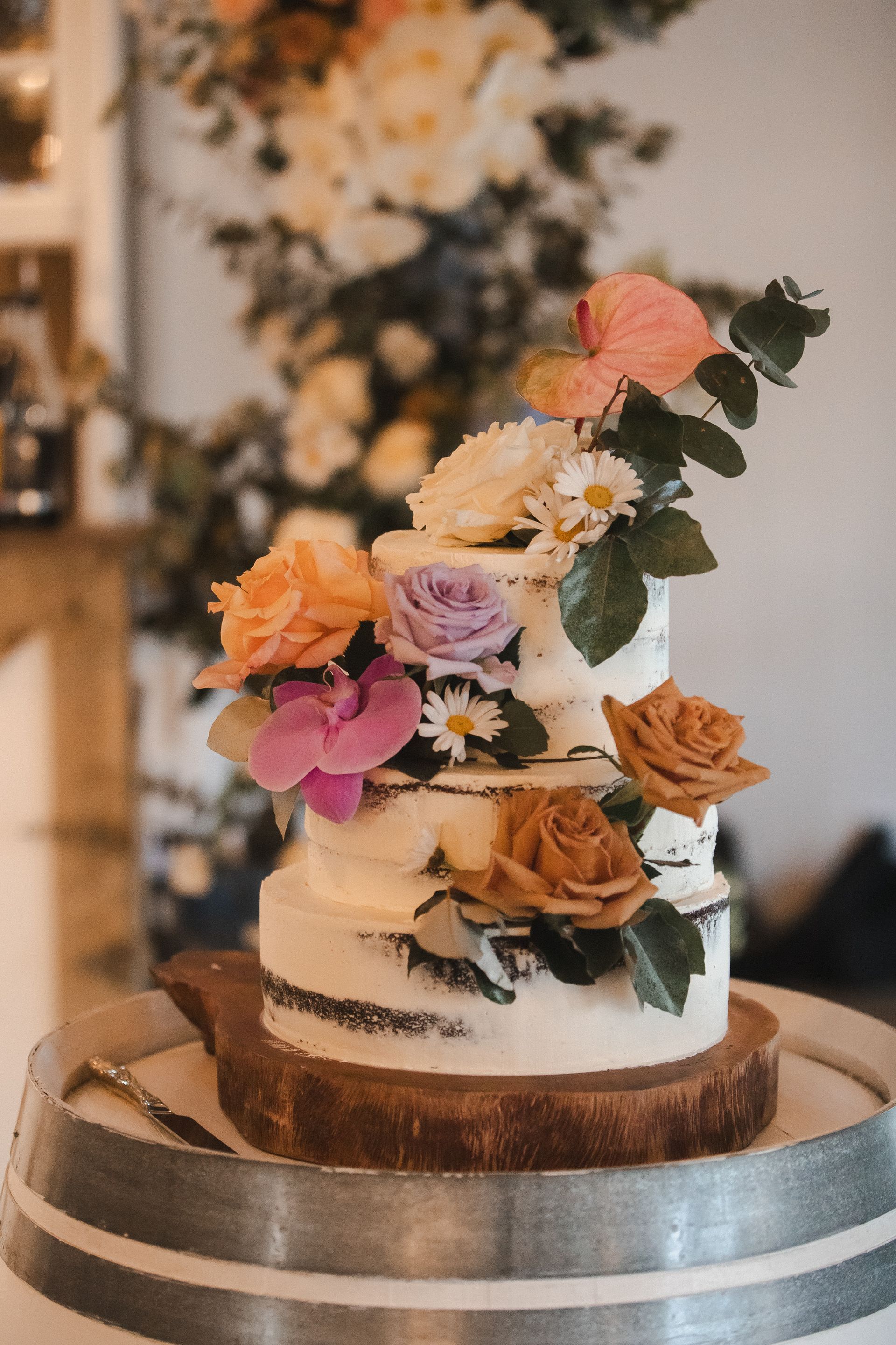 Three-tiered wedding cake decorated with colorful flowers on a wood slice, sitting on a barrel.