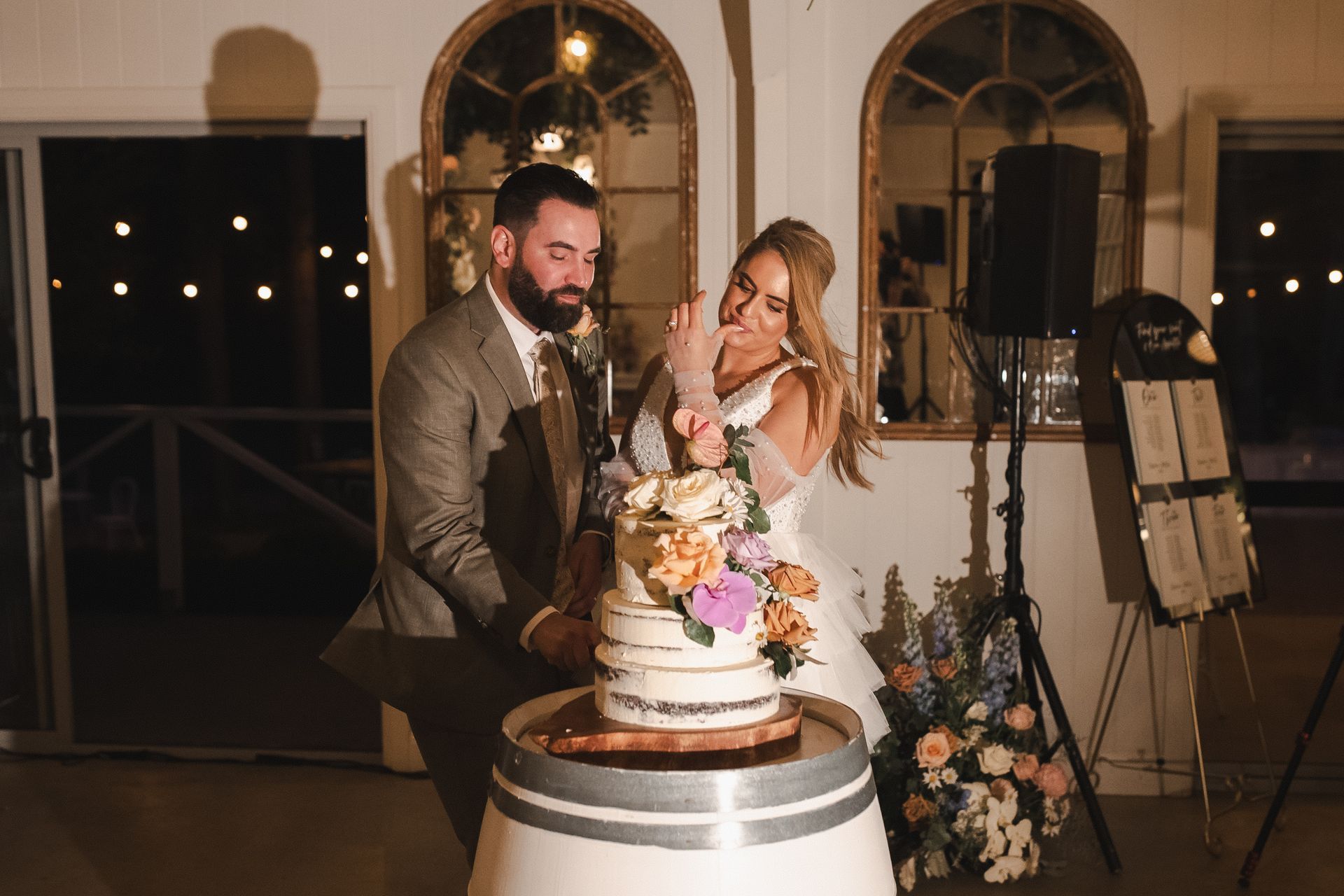 Couple cuts wedding cake, smiling. Outdoors, evening. Cake is floral-adorned on barrel.