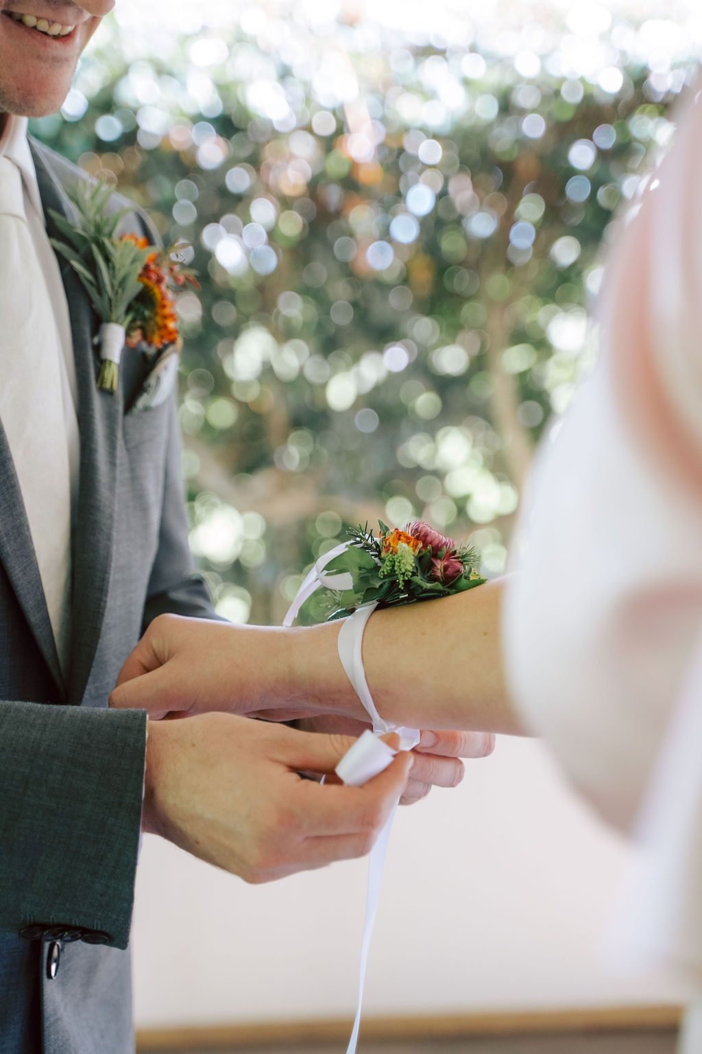 Man ties white ribbon around a person's wrist, wedding ceremony. Green boutonniere, out-of-focus foliage.