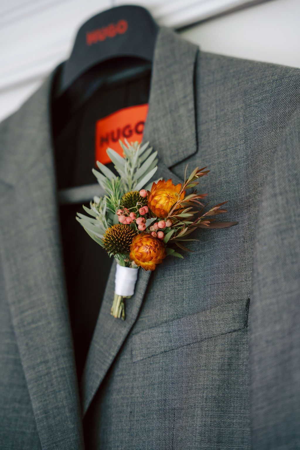 Gray suit jacket with a floral boutonniere pinned to the lapel, hanging on a black HUGO hanger.