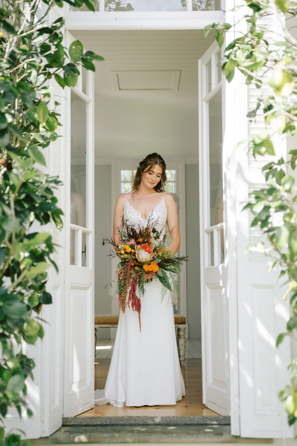 Bride in white dress, holding bouquet, standing in doorway, surrounded by greenery.