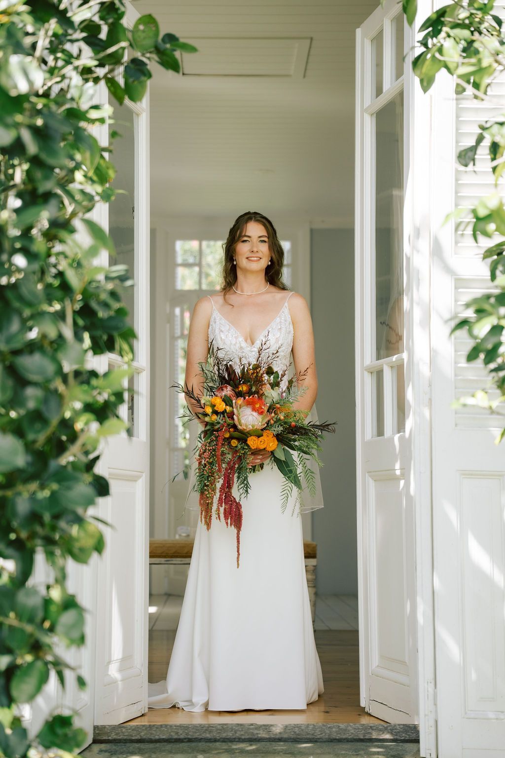 Bride in white dress holding bouquet, standing in doorway, surrounded by greenery.