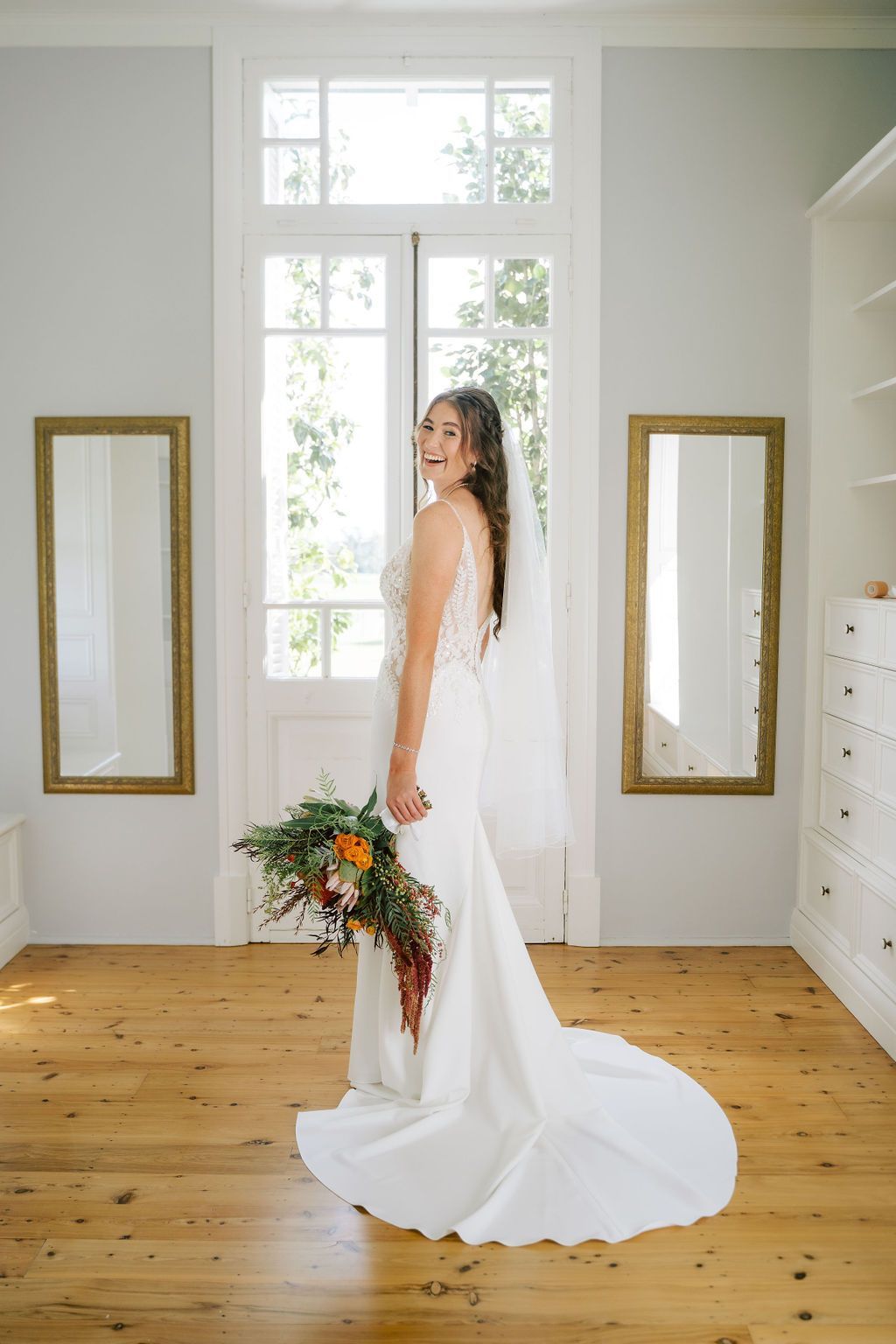 Bride in a white gown holding bouquet, standing by a window with mirrors in a room with wood flooring.