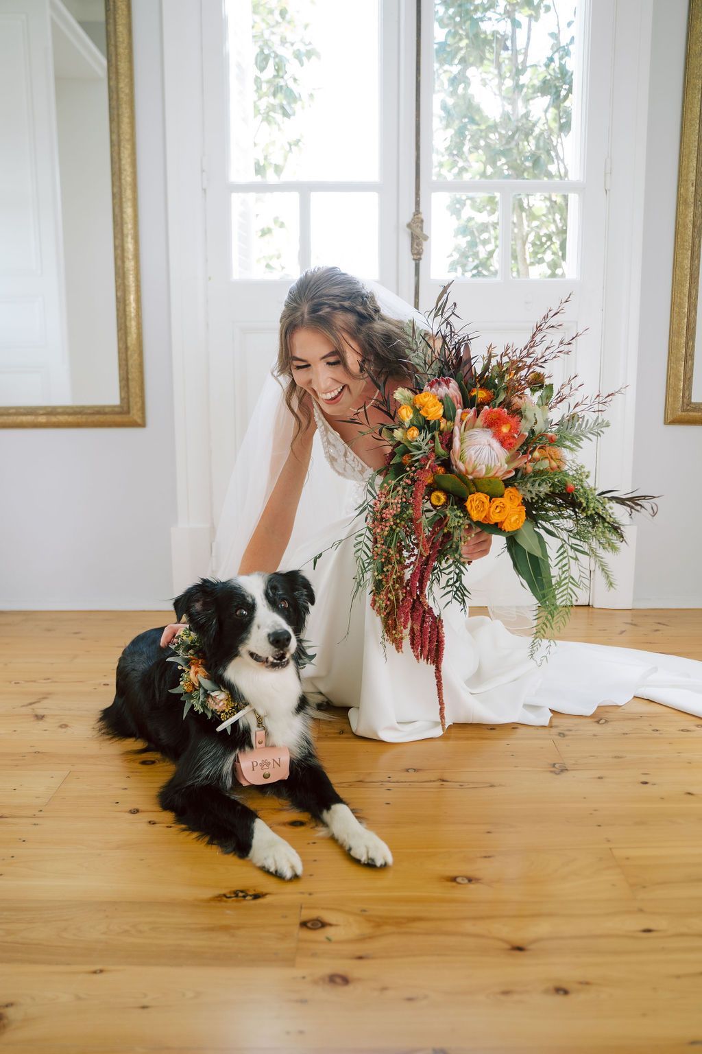 Bride kneels with a dog wearing a floral collar, holding a bouquet. Wooden floor, large window, and mirrors.