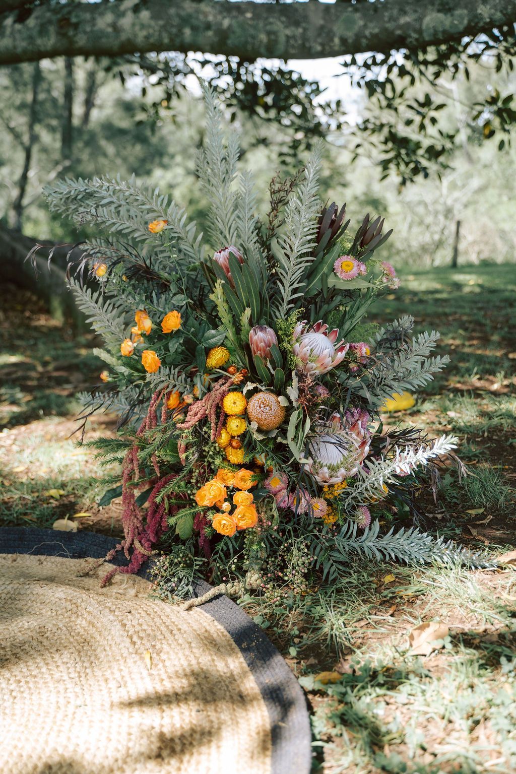 Floral arrangement with protea and orange flowers on a woven mat in a grassy outdoor setting.