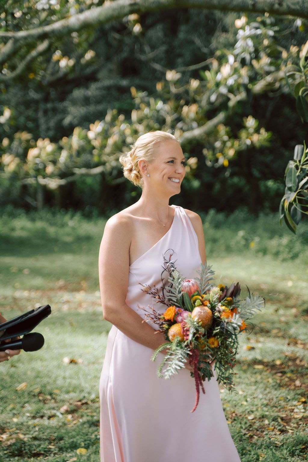 Woman in pink one-shoulder dress holds bouquet, smiles outside.