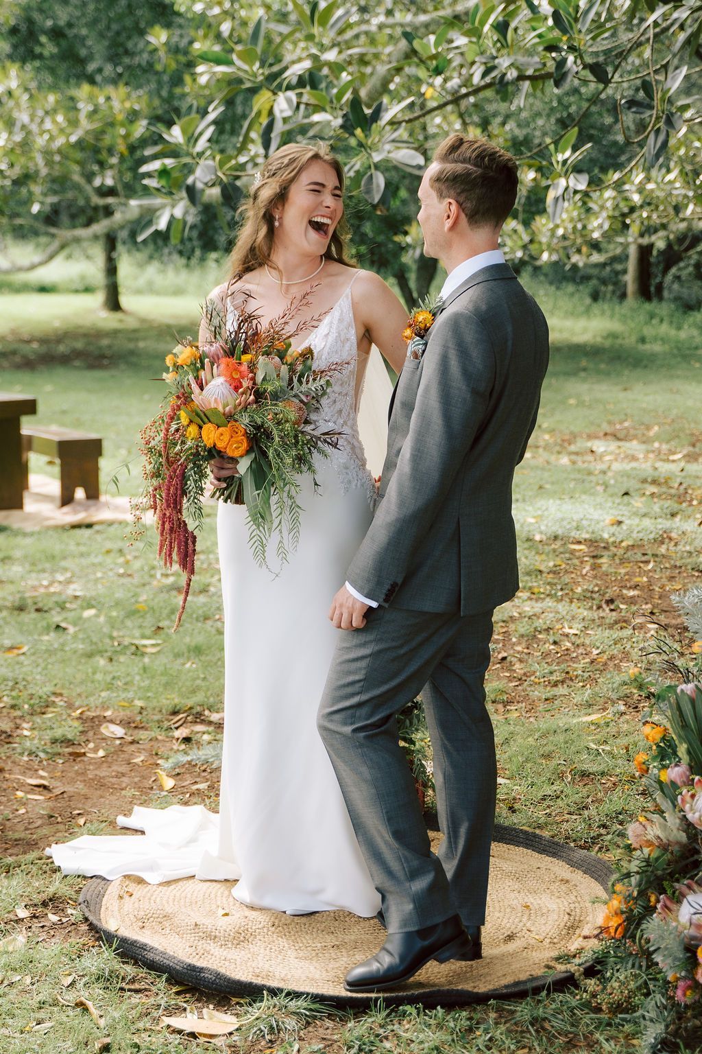 Bride laughs, looking at groom. They stand on a circular mat outdoors with flowers and a tree.