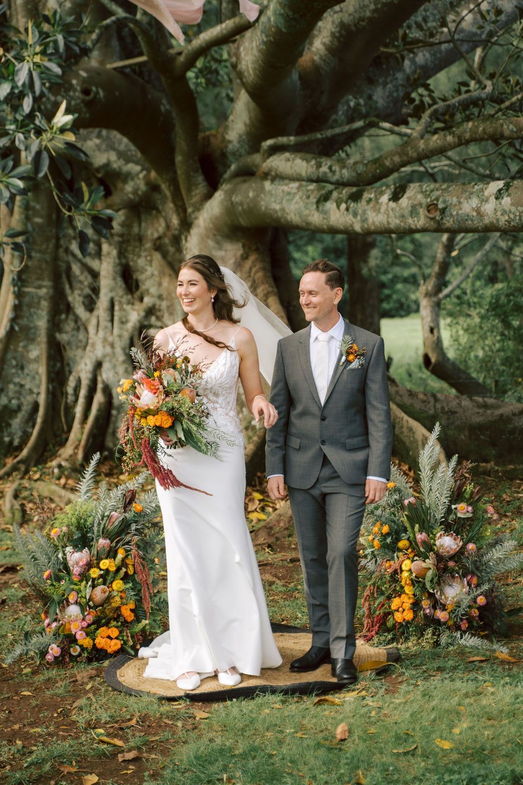 Bride and groom walk hand-in-hand under a large tree during their outdoor wedding ceremony. 