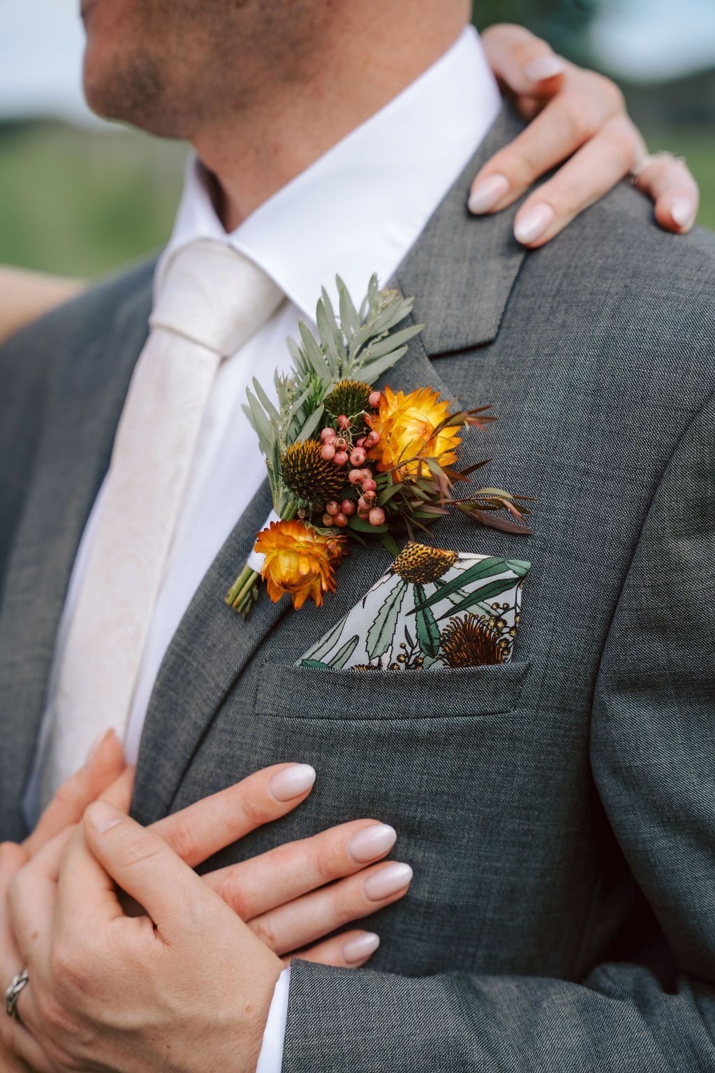 Man in gray suit with boutonniere and pocket square, embraced by someone's hands.