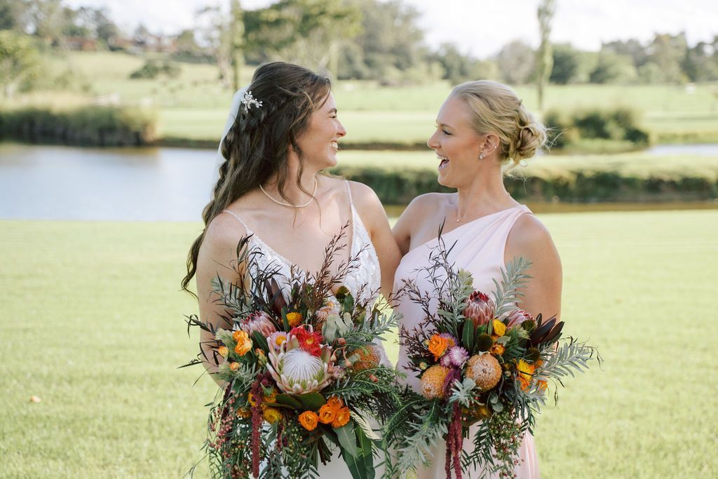 Two women smiling, holding bouquets in a grassy field near water.