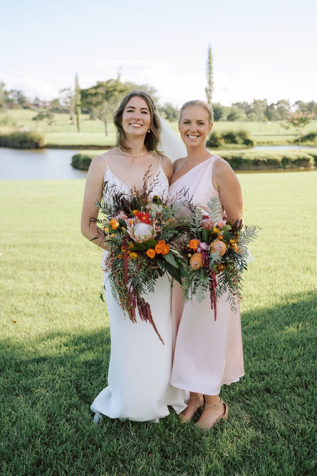 Bride and bridesmaid pose outdoors, holding bouquets. The bride wears white, bridesmaid pink. Sunny, green field.