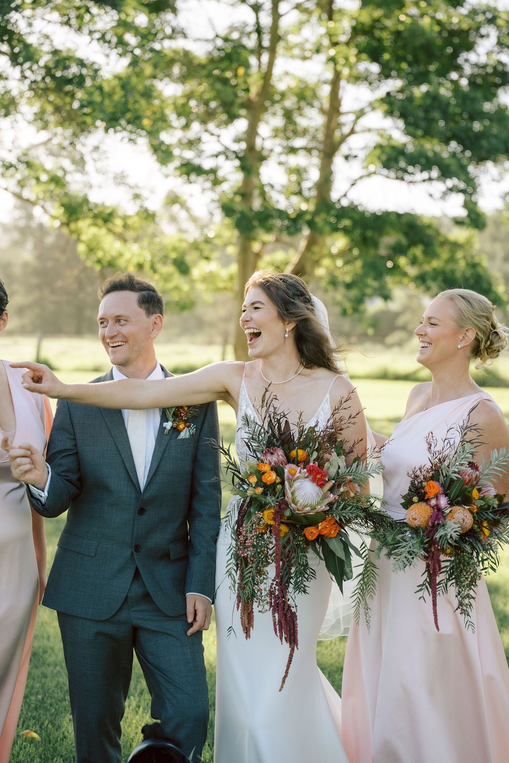 Wedding party laughing in a field. Bride in white dress points; groom smiles. Bridesmaid in pink holds bouquet.