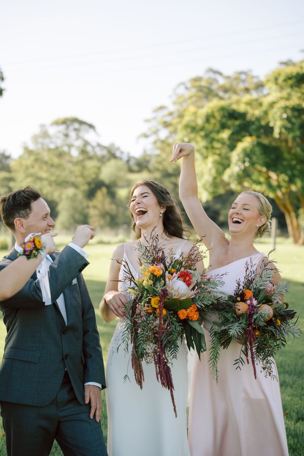 Wedding party cheers outdoors: bride and bridesmaid laughing with bouquets, groomsman in suit raises fist.