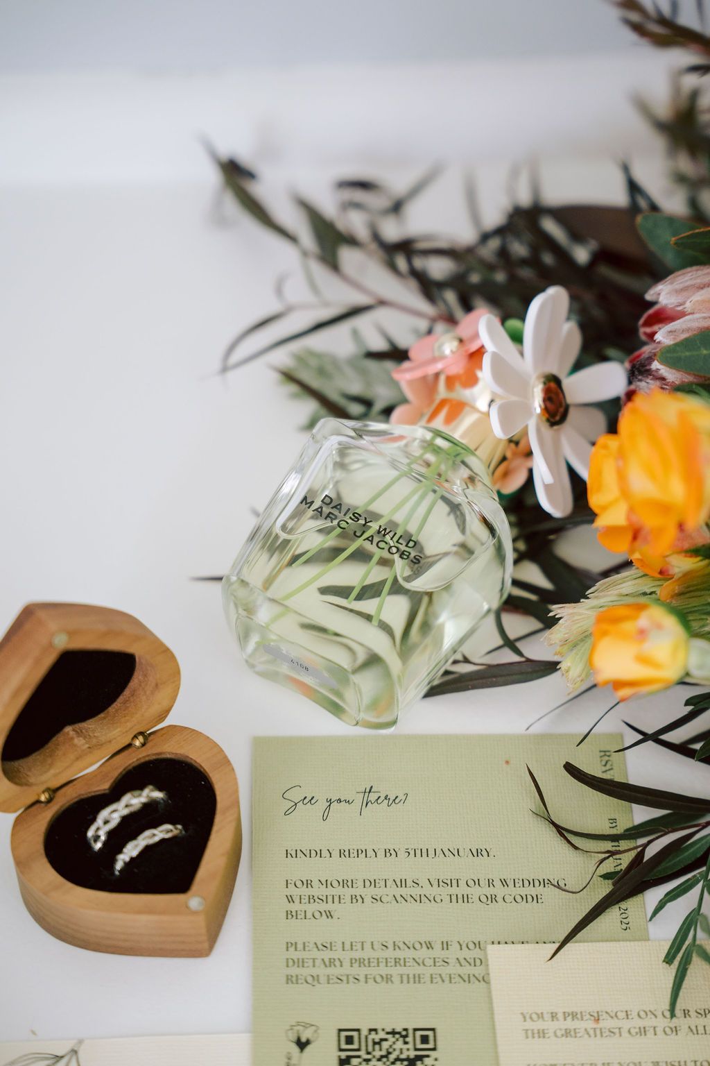 Wedding rings in a heart-shaped box, floral arrangement, glass perfume bottle, and invitation on white surface.