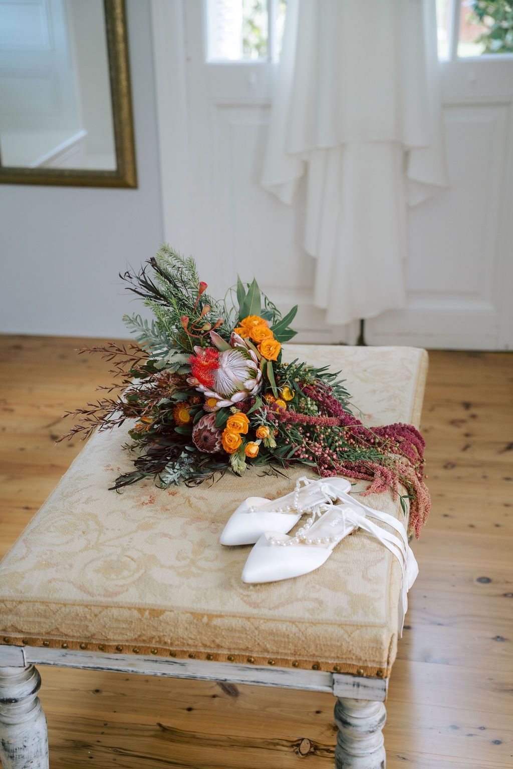Wedding bouquet, shoes, and dress on a cushioned bench in a bright room.