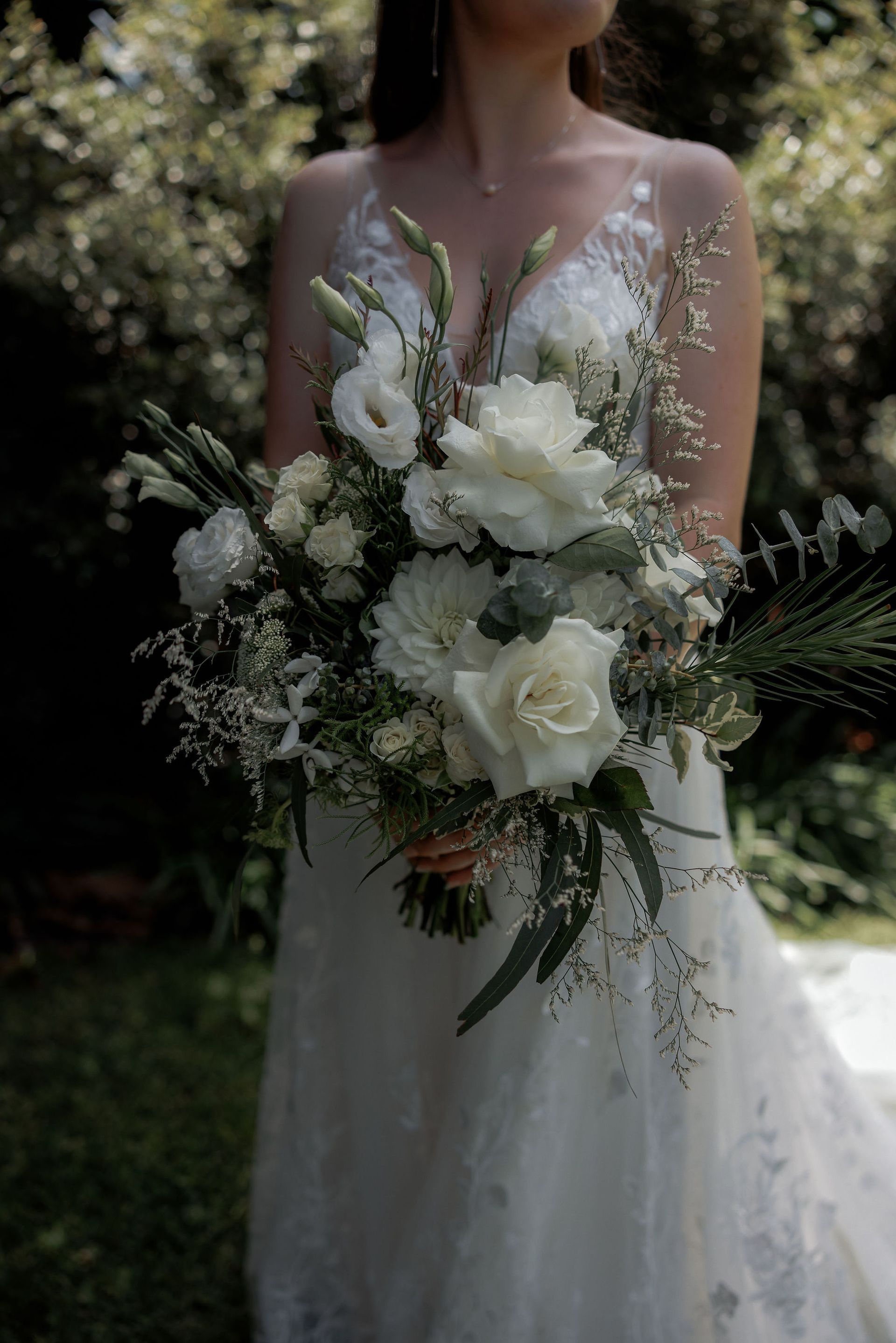 Bride in white lace dress holding a bouquet of white flowers outdoors in sunlight.