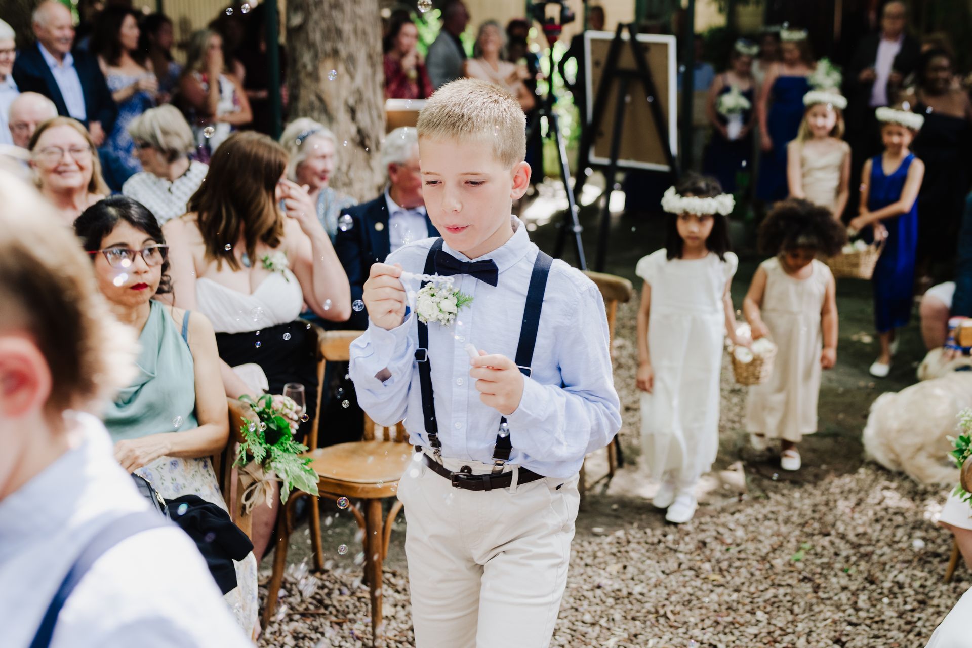 Boy in bow tie walking down aisle at a wedding, confetti falling. Flower girls follow.