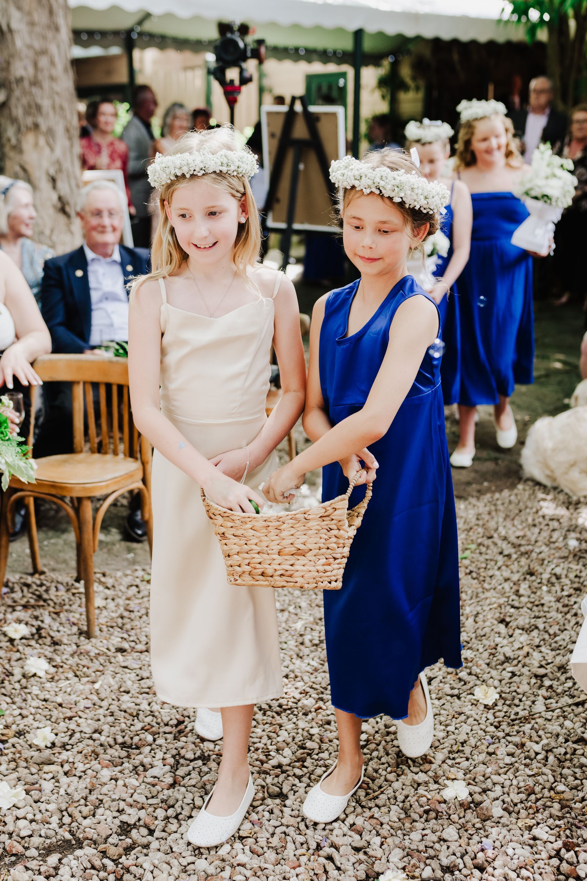 Two young girls in floral crowns carry a basket, followed by a bridesmaid in a blue dress at an outdoor wedding.
