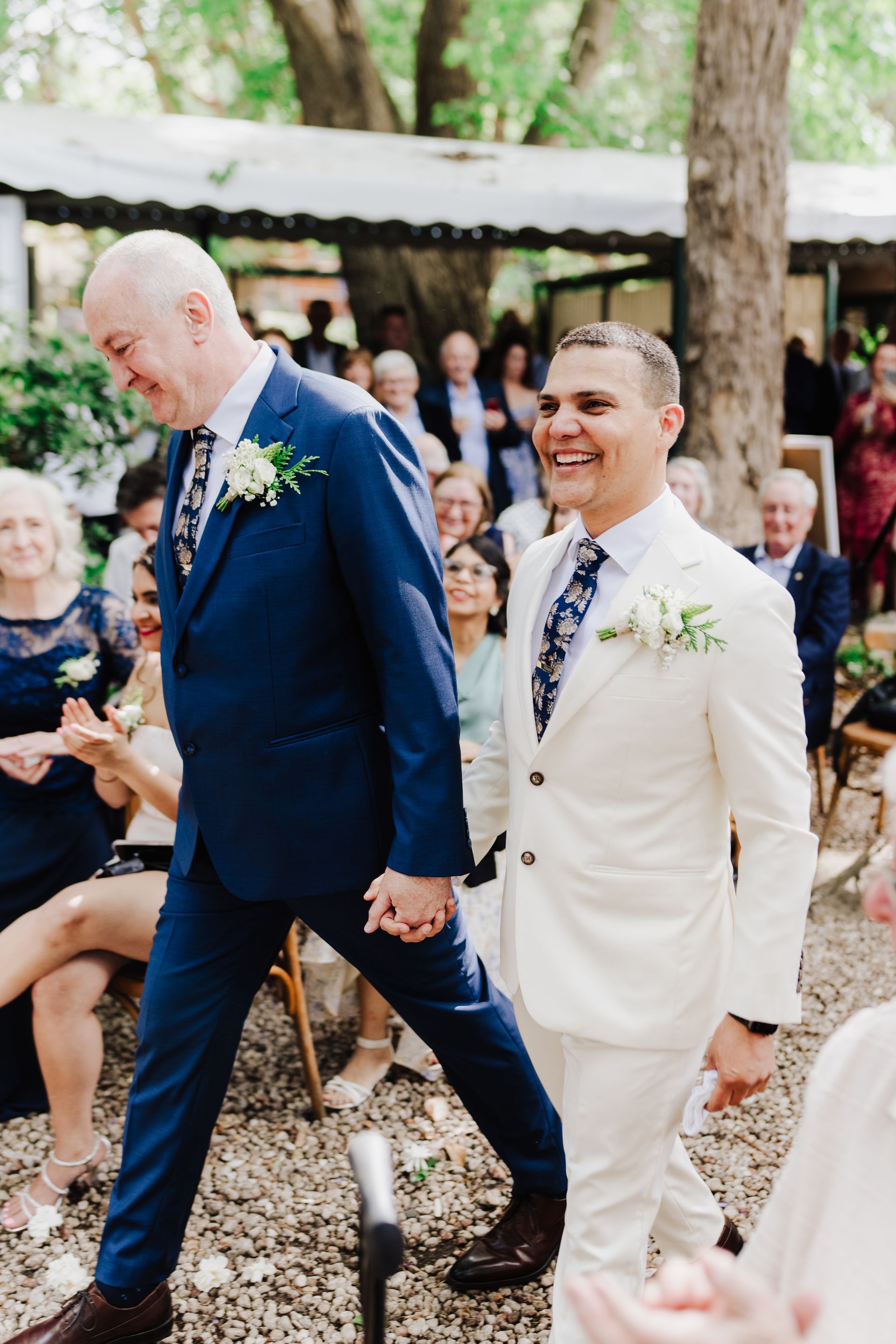 Two grooms, one in blue suit, the other in white, smiling, holding hands, walking down aisle at outdoor wedding.